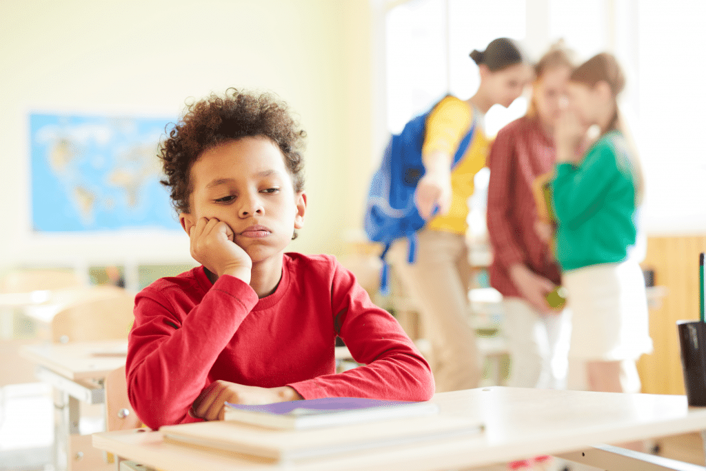 A child in red clothing sitting alone in the classroom while girls whispering and pointing at him