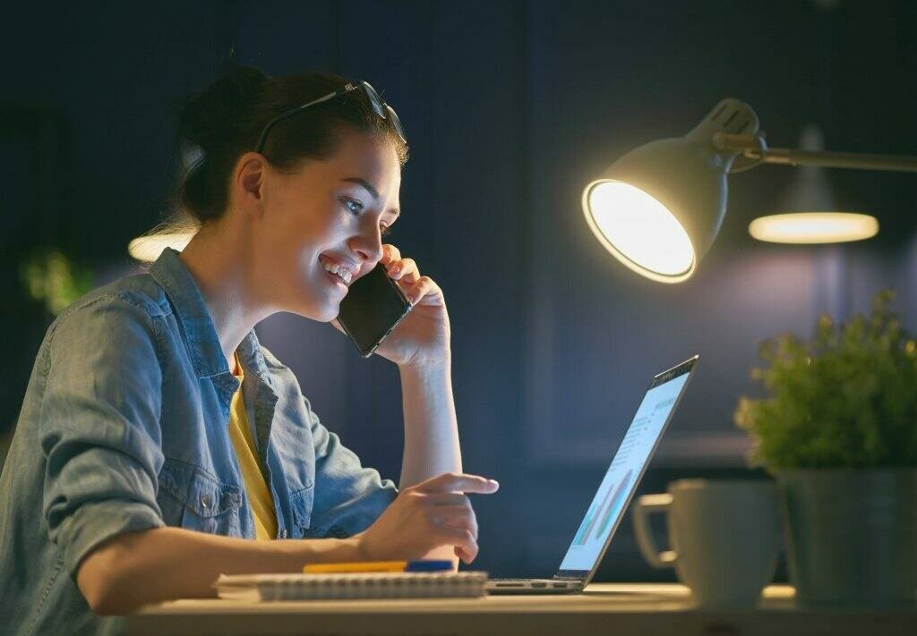 Smiling woman working at night and on a phone call while using her laptop